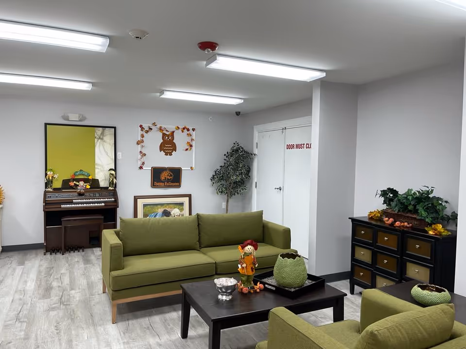 A cozy living room area with green sofas and armchairs arranged around a dark wooden coffee table. The table has decorative items including a small scarecrow figure and a green textured vase. In the background, there is a piano with small decorations on top, a large mirror, a framed picture leaning against the wall, and a whiteboard with a Halloween-themed decoration. A potted plant is placed near a white double door with a sign that reads 'DOOR MUST CL'. The room has light gray walls and wood-patterned flooring, illuminated by ceiling fluorescent lights.