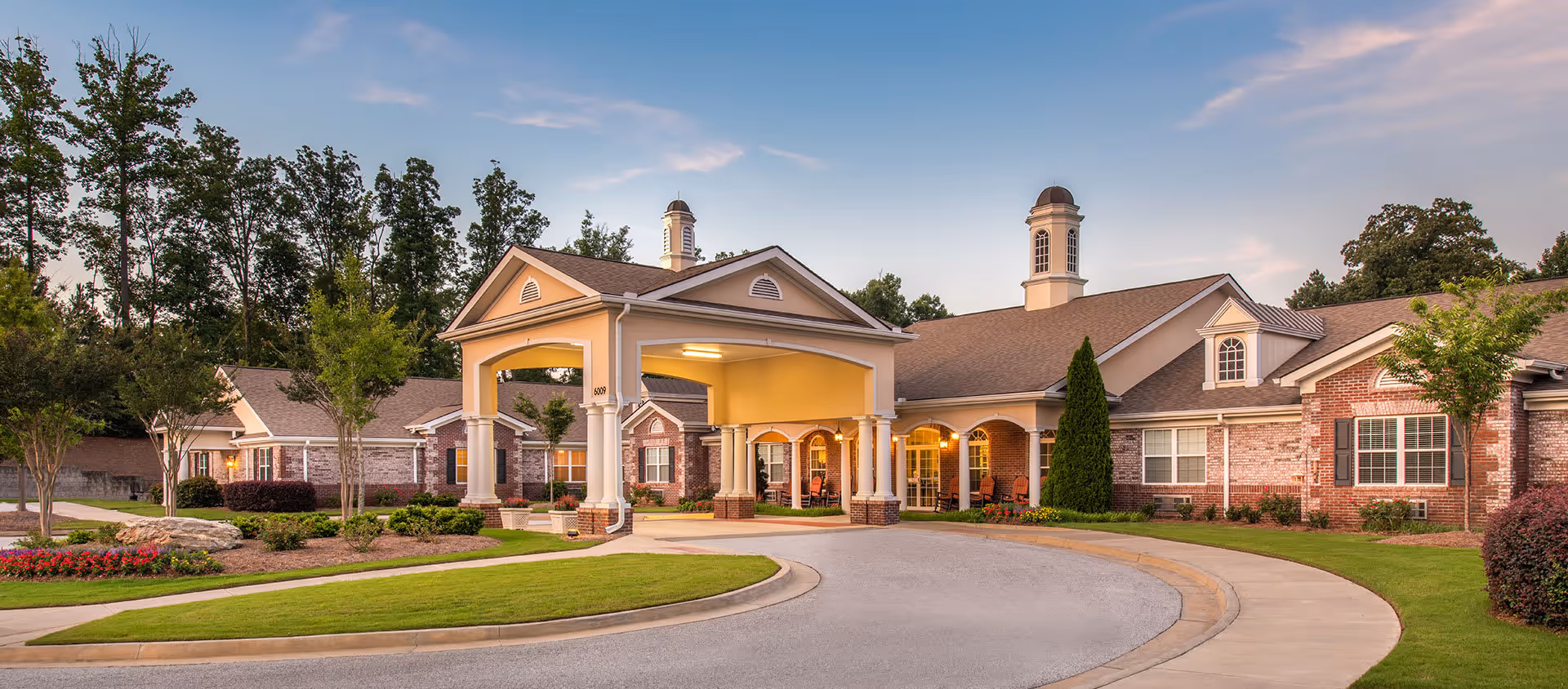 Front entrance of a single-story brick senior living facility with a porte-cochère, circular driveway and landscaped grounds at dusk.