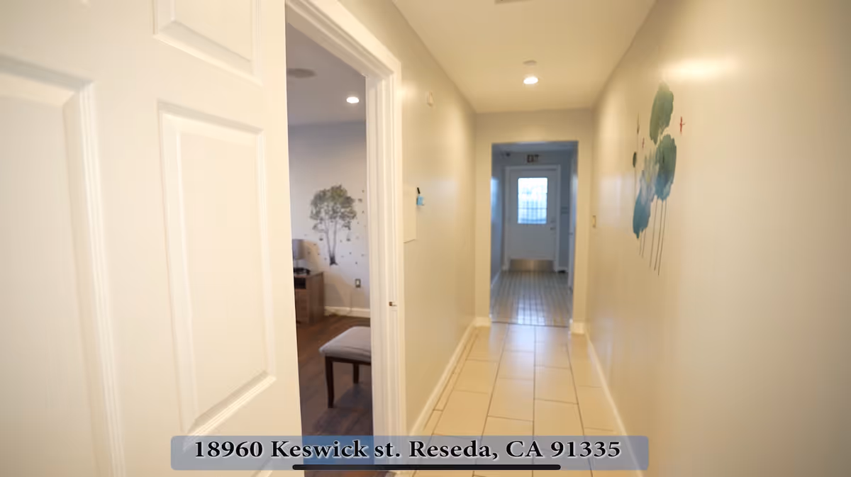 A clean, well-lit hallway in an assisted living facility with beige walls and tiled floor. On the left, there is an open door leading to a room with wooden flooring and a wall decal of a tree. The hallway leads to a door with a window at the far end. There is a floral wall decoration on the right wall of the hallway.