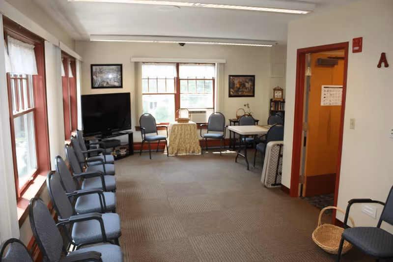 A well-lit common room with multiple chairs arranged along the left wall and around tables near the windows. There is a flat-screen TV on a stand in the corner, framed pictures on the walls, and a basket on a chair in the foreground. The room has large windows with white curtains and red trim, beige walls, and a carpeted floor.