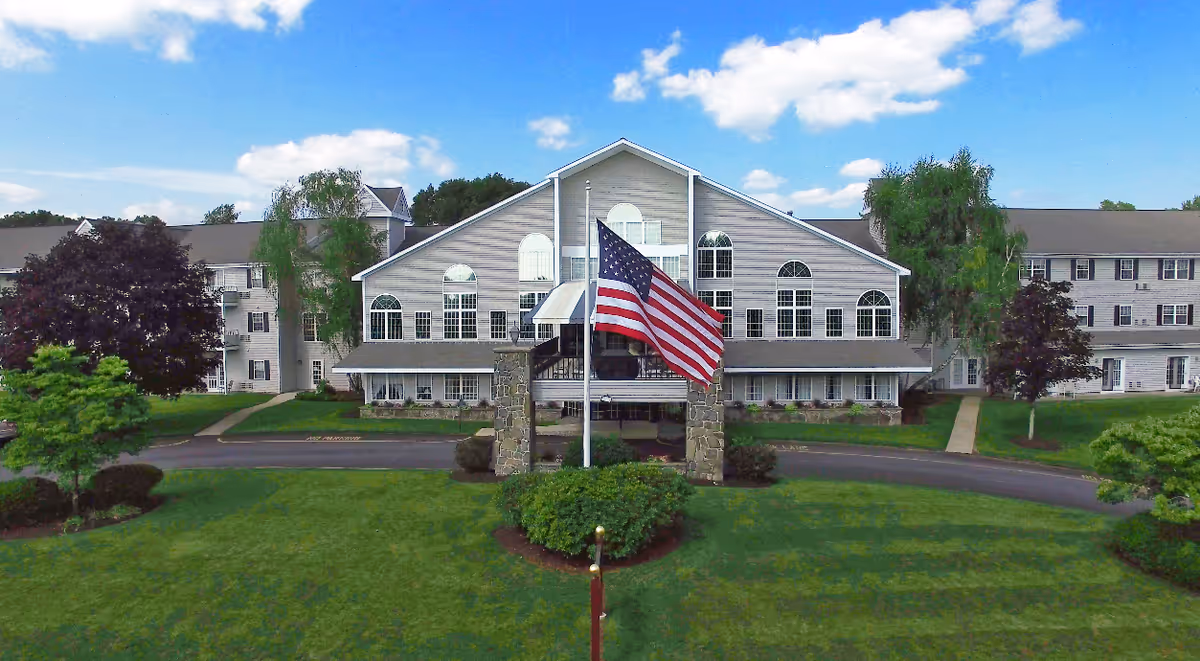 Front exterior view of a large senior living facility building with multiple windows and a peaked roof. An American flag is prominently displayed on a flagpole in the center of a circular driveway surrounded by green lawns and trees under a partly cloudy blue sky.