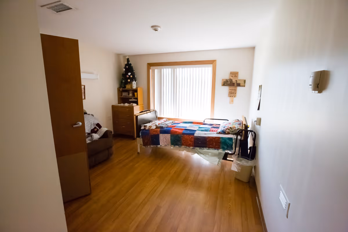 A memory care bedroom with a single bed, patchwork quilt, dresser, and large window.