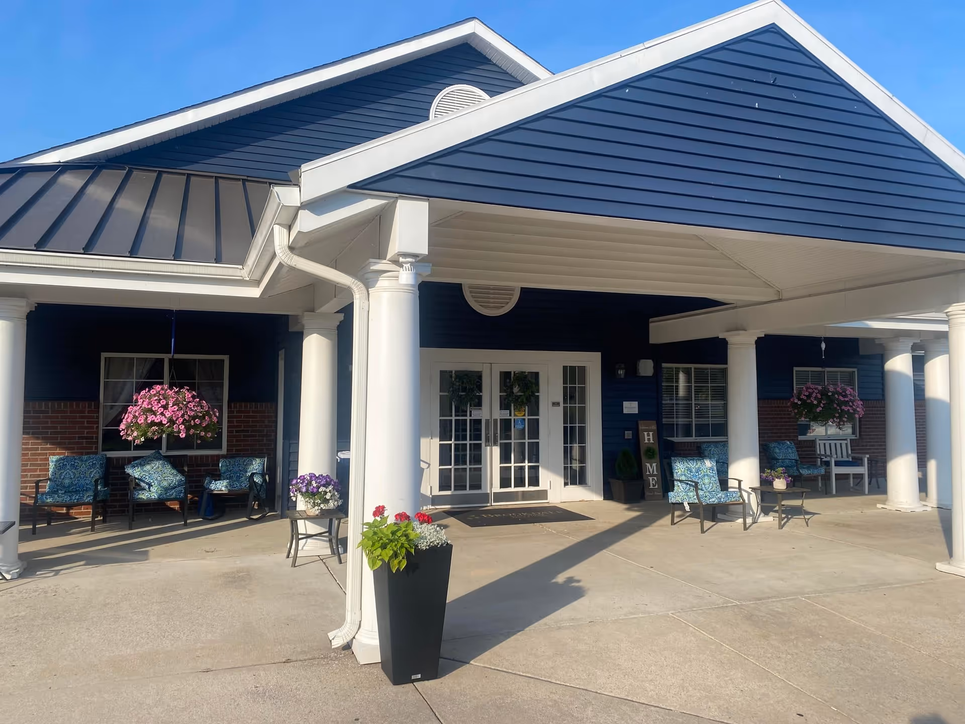 Entrance of a building with a covered porch supported by white columns. There are several chairs with blue patterned cushions and hanging flower baskets on either side of the entrance. The building has dark blue siding with white trim and a brick lower facade. A black planter with flowers is placed near one of the columns.
