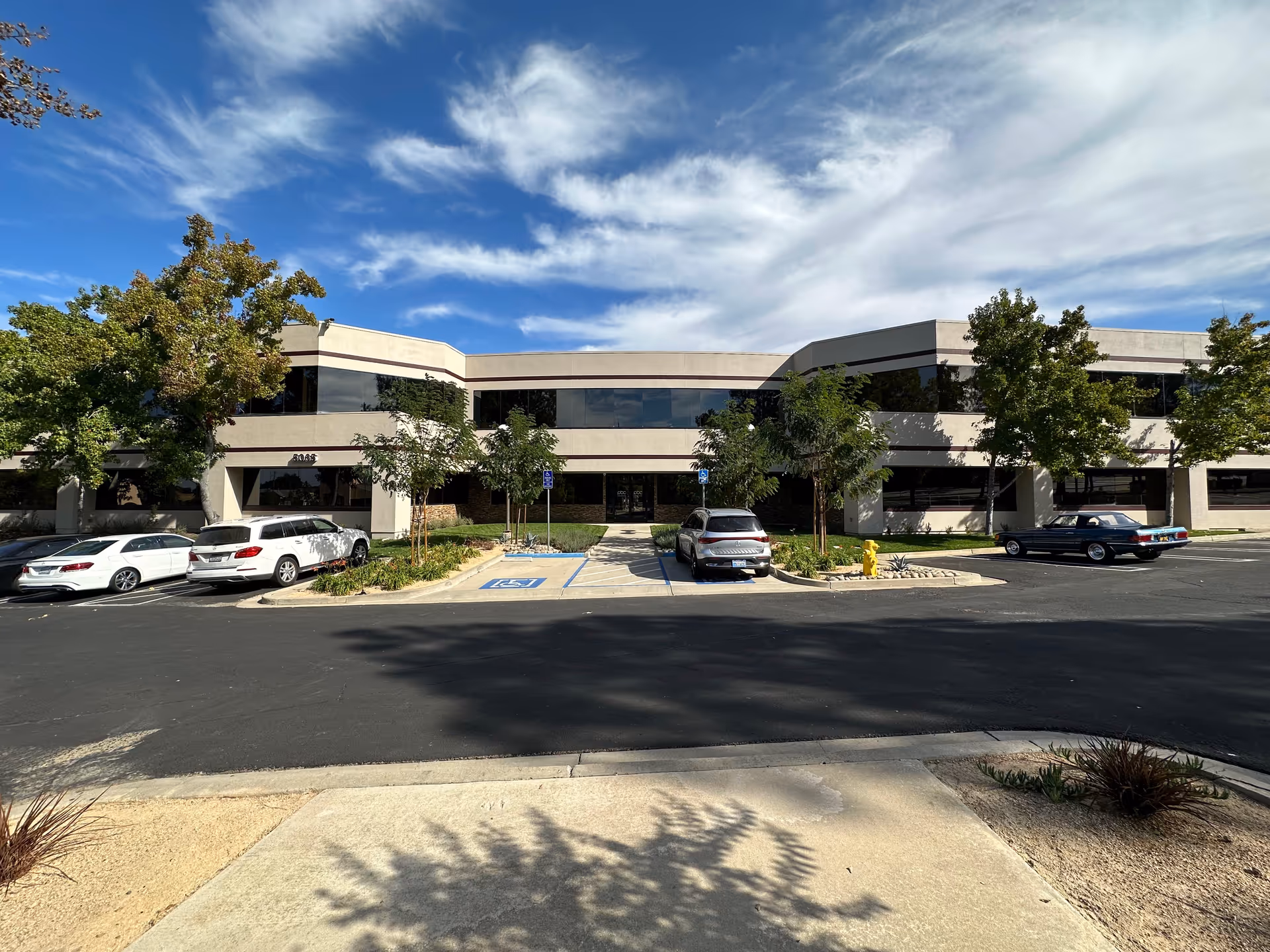 Two-story beige office building with parked cars and trees in a sunny parking lot.