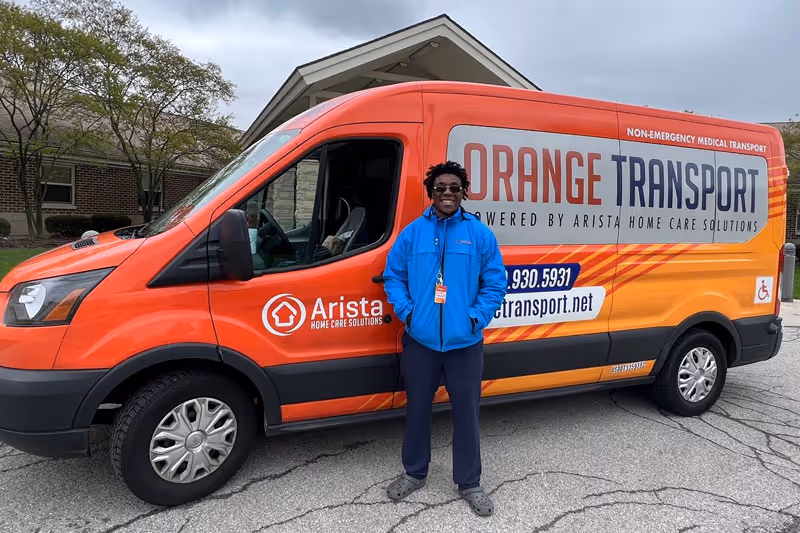 A staff member stands beside an Orange Transport van branded Arista Home Care Solutions.