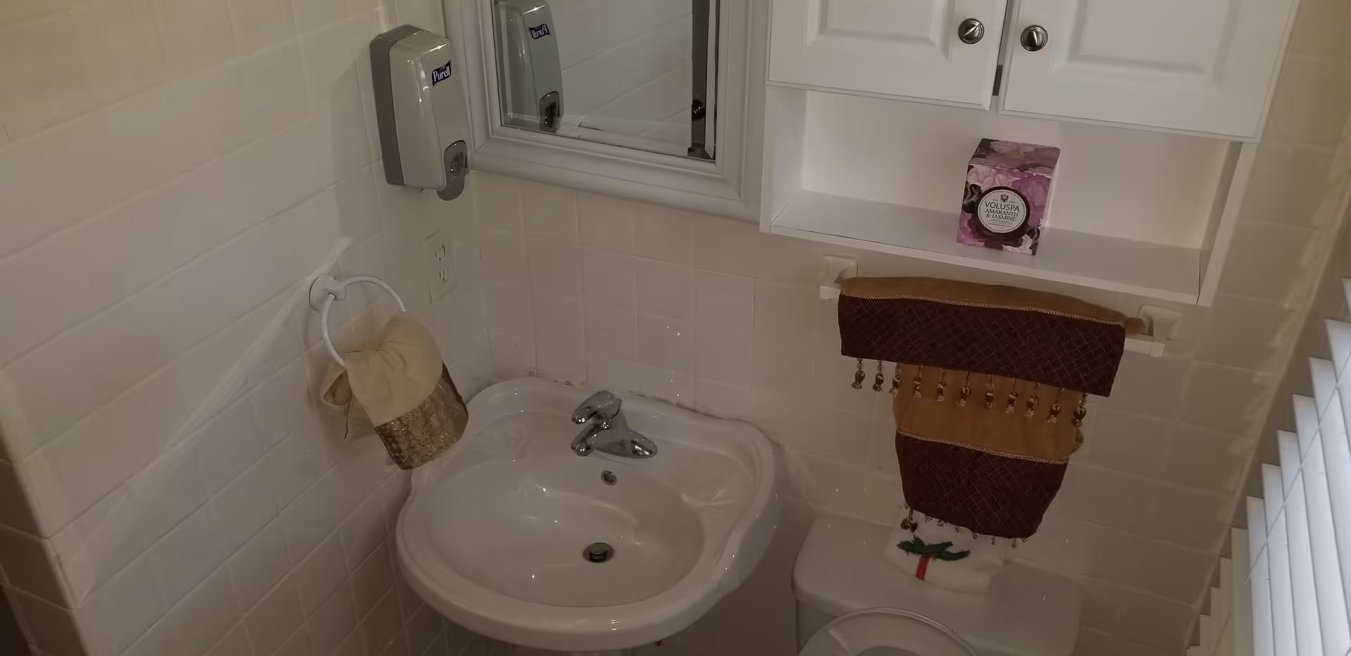 Small bathroom with white tiled walls featuring a white sink with a silver faucet, a mirror above the sink, a soap dispenser mounted on the wall, a towel ring with a beige and brown hand towel, a white cabinet with two doors above the toilet, and a brown and beige decorative towel hanging on a towel bar above the toilet.