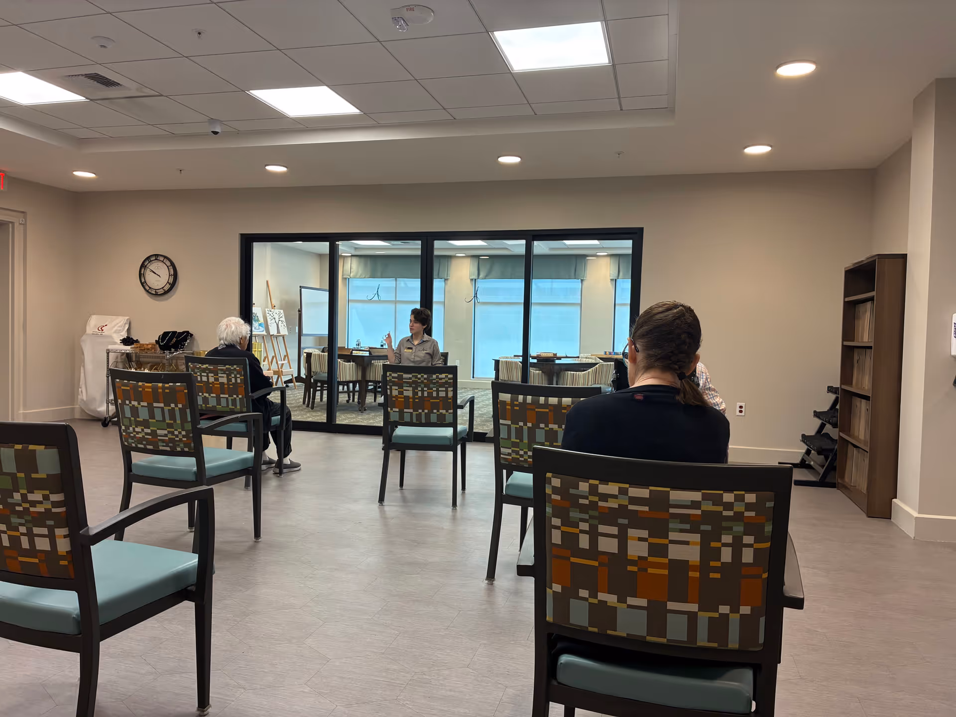 Interior waiting area with patterned chairs, residents seated, and staff behind a glass partition.