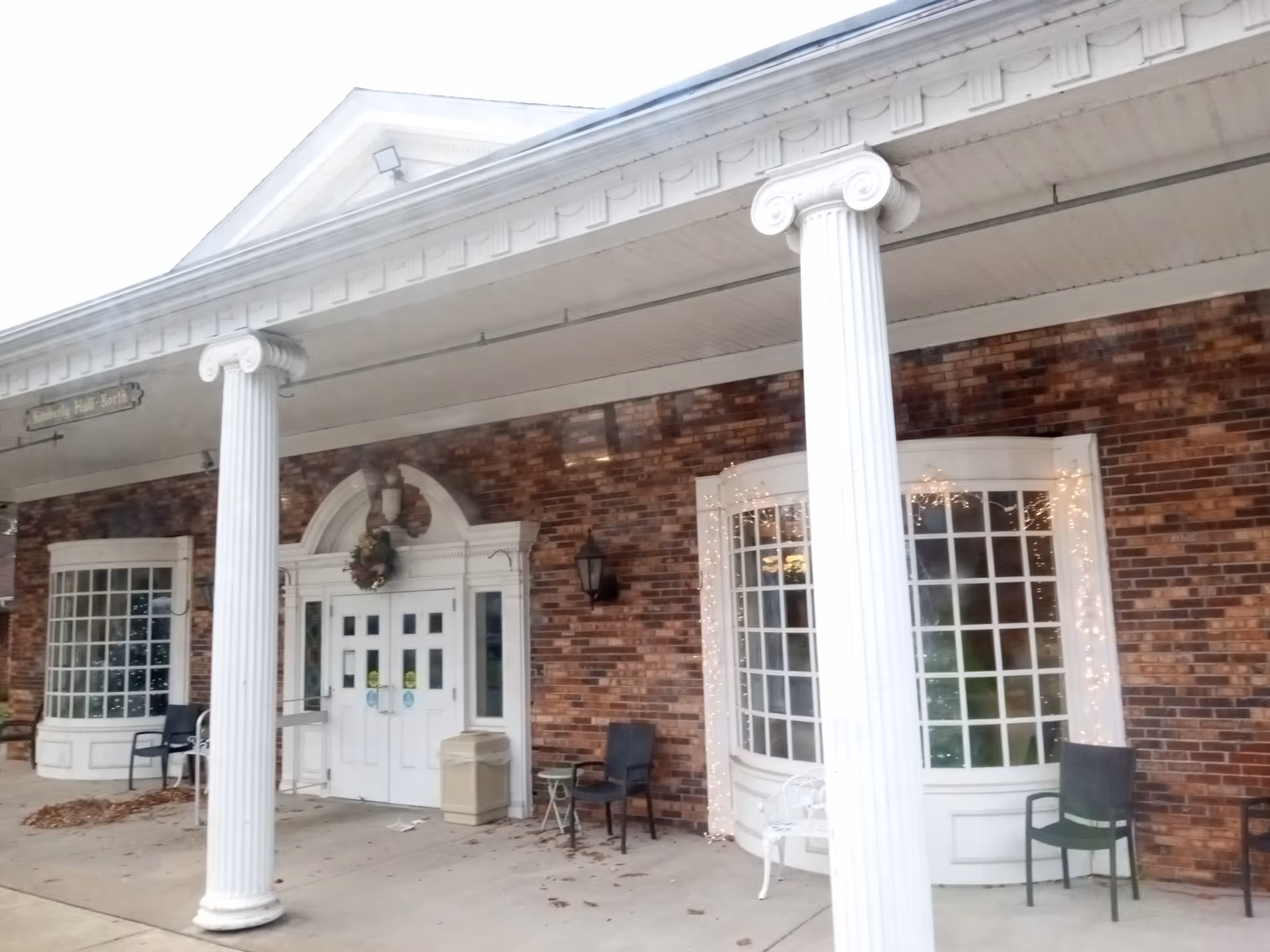 Brick building entrance with white columns, double doors, wreath, and outdoor seating.