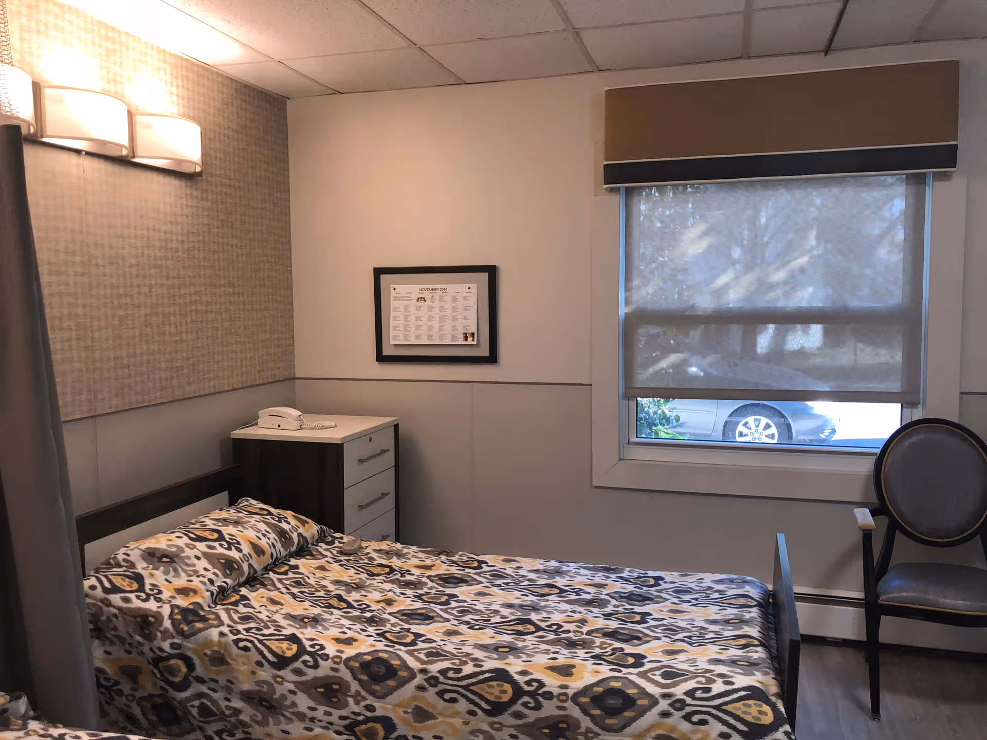 A furnished senior living bedroom with patterned bedspread, window, and nearby chair.