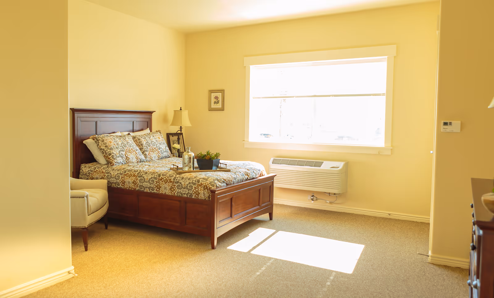 Furnished bedroom with a wooden bed dressed in patterned bedding, an armchair, a bedside lamp and a large sunlit window.