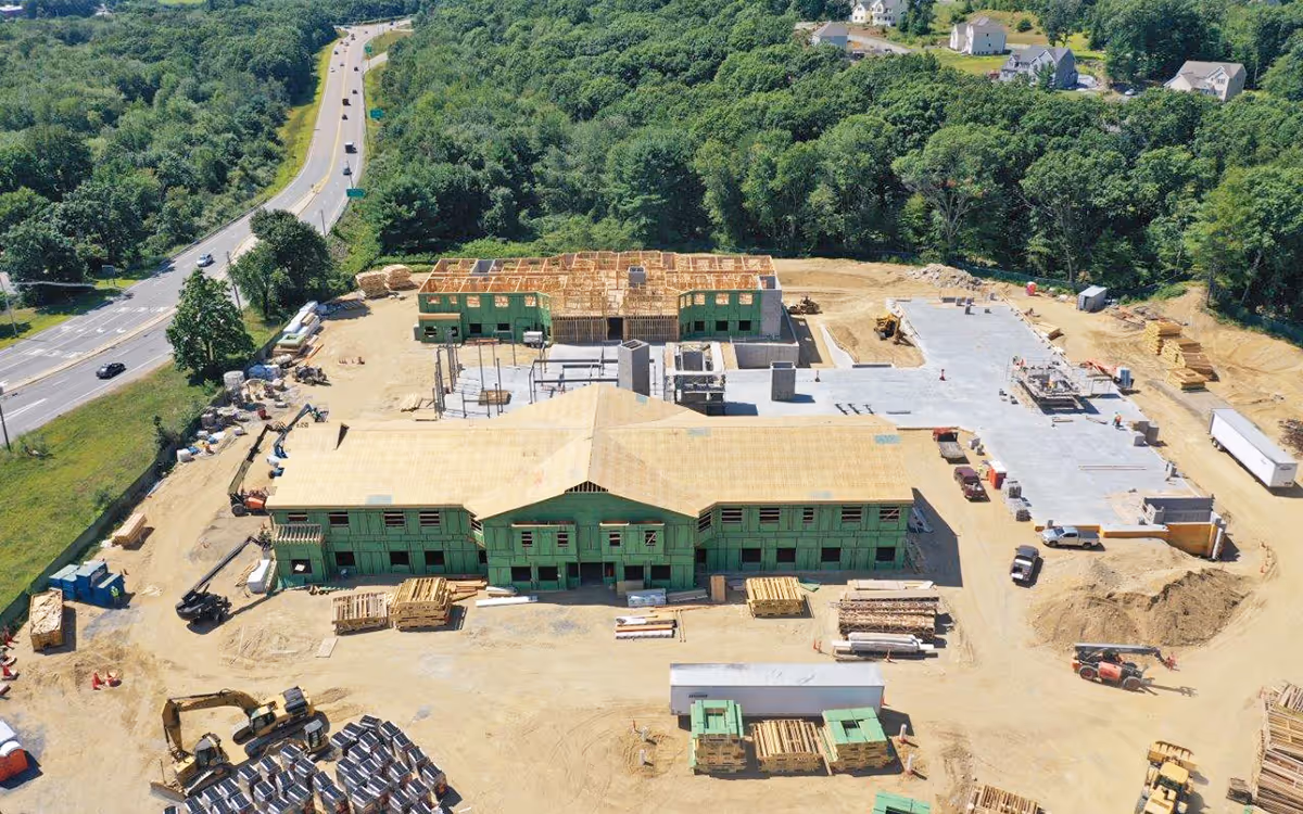 Aerial view of the Artisan at Hudson construction site showing a large two-story building with green sheathing, construction equipment, vehicles, and cleared land surrounded by trees and a nearby road.