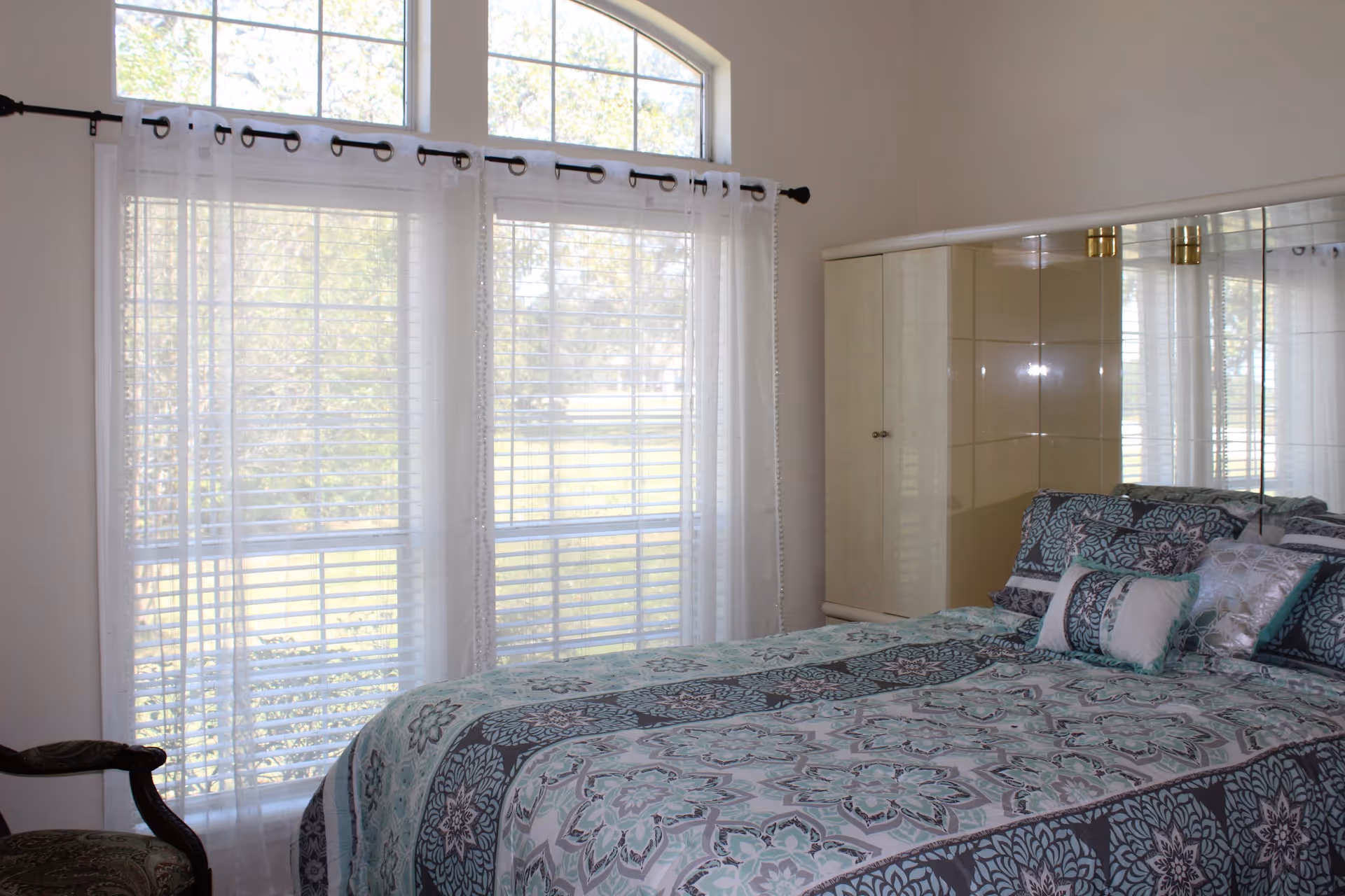 Sunlit bedroom featuring a patterned bed, mirrored wardrobe, and large windows with blinds and sheer curtains.