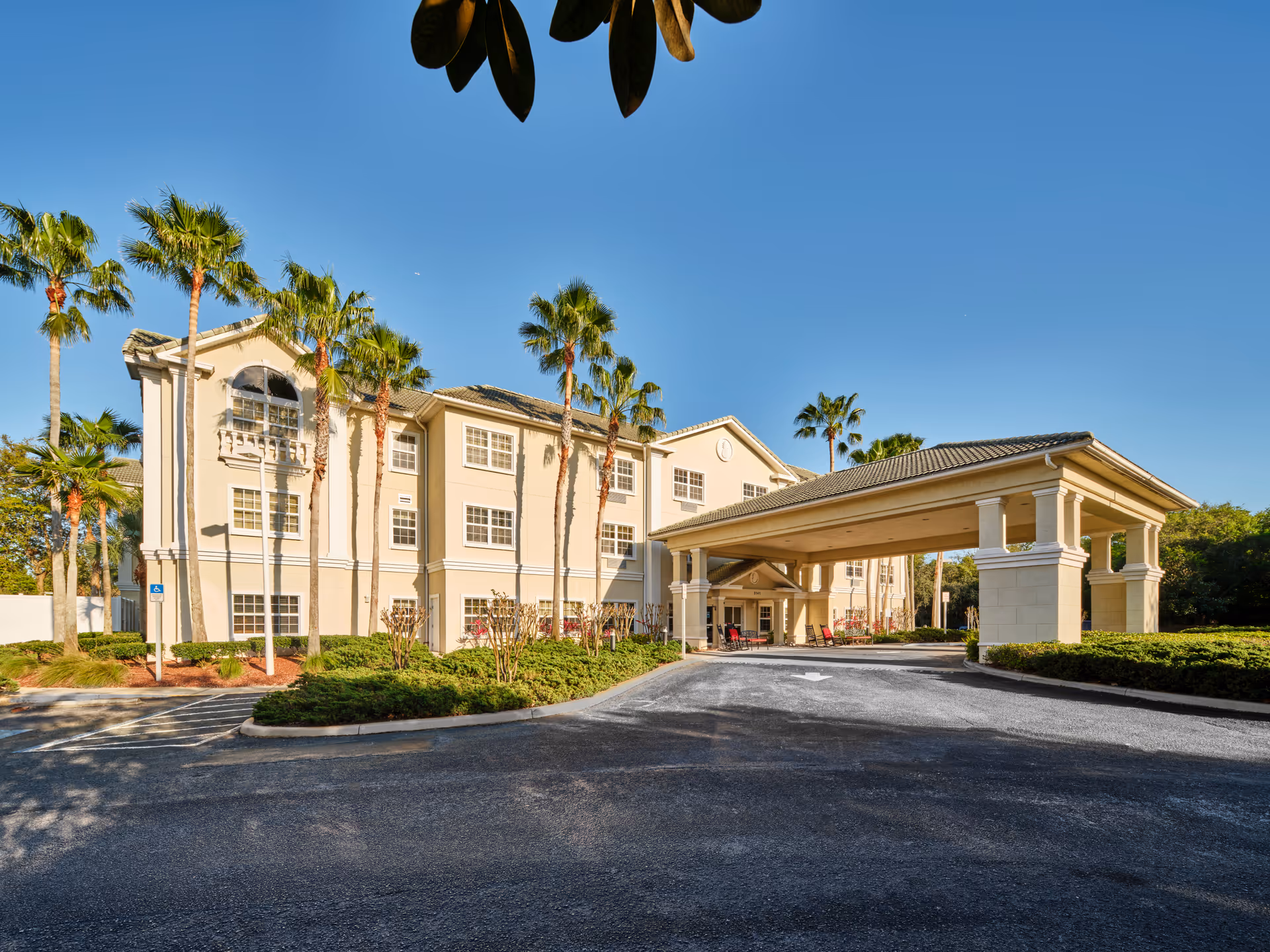 Front exterior of a three-story beige assisted living building with a covered porte-cochere, palm trees, and a clear blue sky.