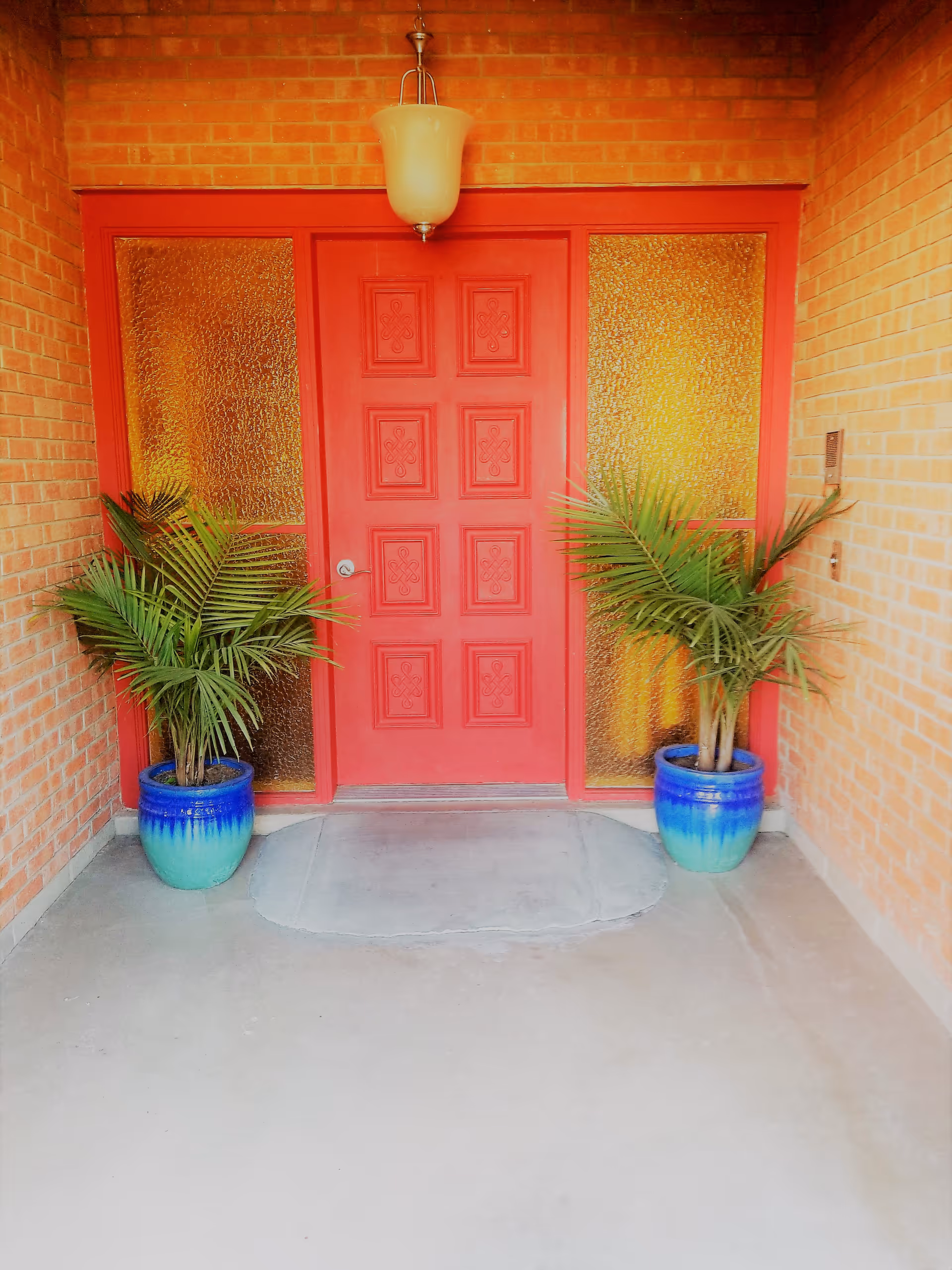 Entrance area with a red door featuring decorative panels, flanked by two textured glass side panels. Two potted green plants in blue gradient pots are placed symmetrically on either side of the door. The walls are made of red brick, and a hanging light fixture is centered above the door.