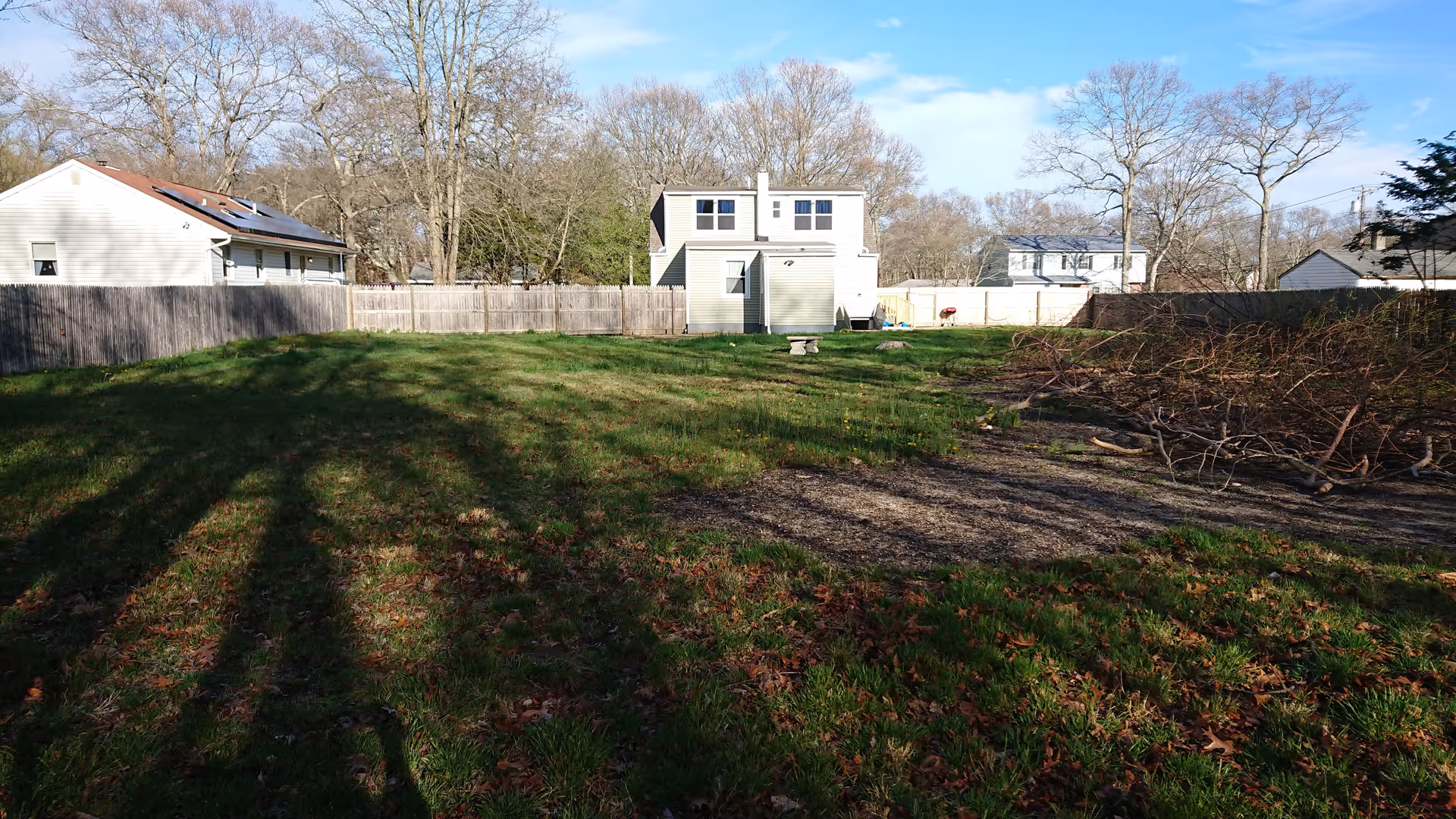 Grassy backyard with wooden fence, a two-story building, leafless trees, and a pile of branches.