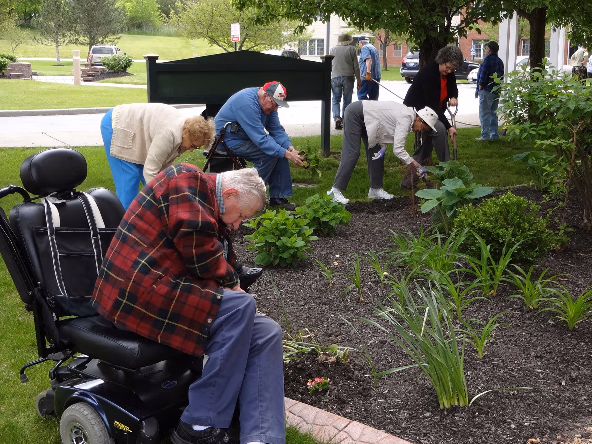 A group of elderly people gardening together in a landscaped outdoor bed, with a man in a motorized wheelchair in the foreground.