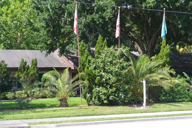 Front exterior of a senior living facility with flags, trees, and manicured shrubs.