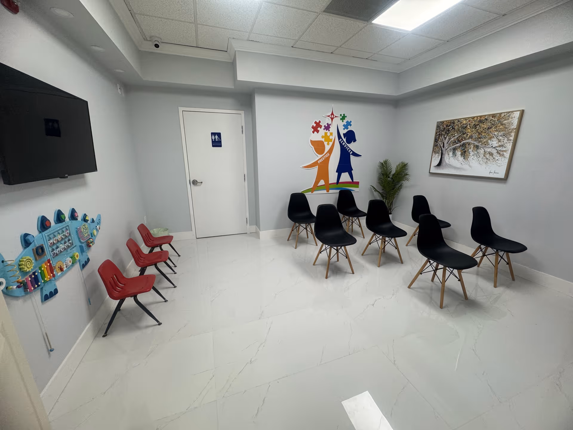 Bright waiting area with black and red chairs, wall art, a plant, and a restroom door.