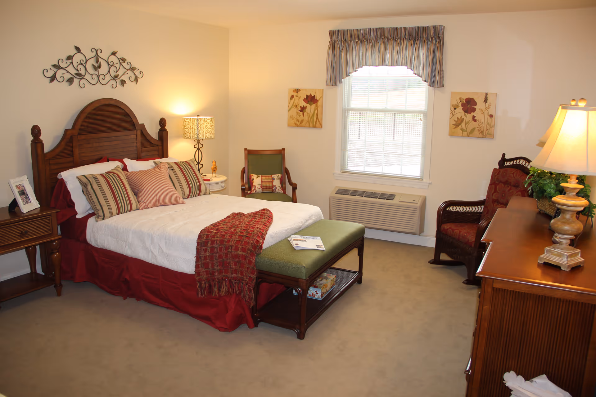 A cozy bedroom with a wooden headboard bed dressed in white and red bedding, multiple striped and solid pillows, and a red throw blanket. There is a green cushioned bench at the foot of the bed with a magazine on it. The room has beige walls, a window with blinds and a striped valance, two floral paintings on the wall, a wooden nightstand with a lamp and picture frame, a green armchair, a red cushioned chair, and a wooden dresser with a lamp and a plant.