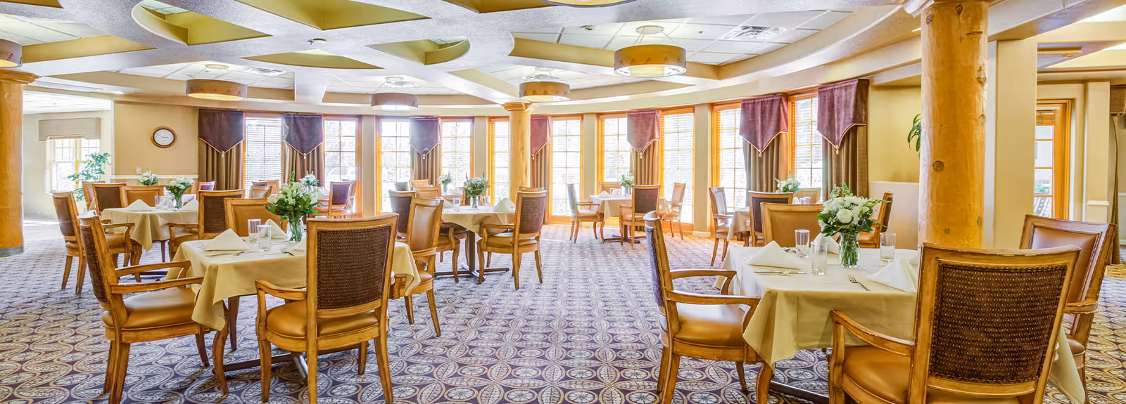Bright, carpeted dining room with multiple tables set with tablecloths, chairs, and floral centerpieces in front of large windows.