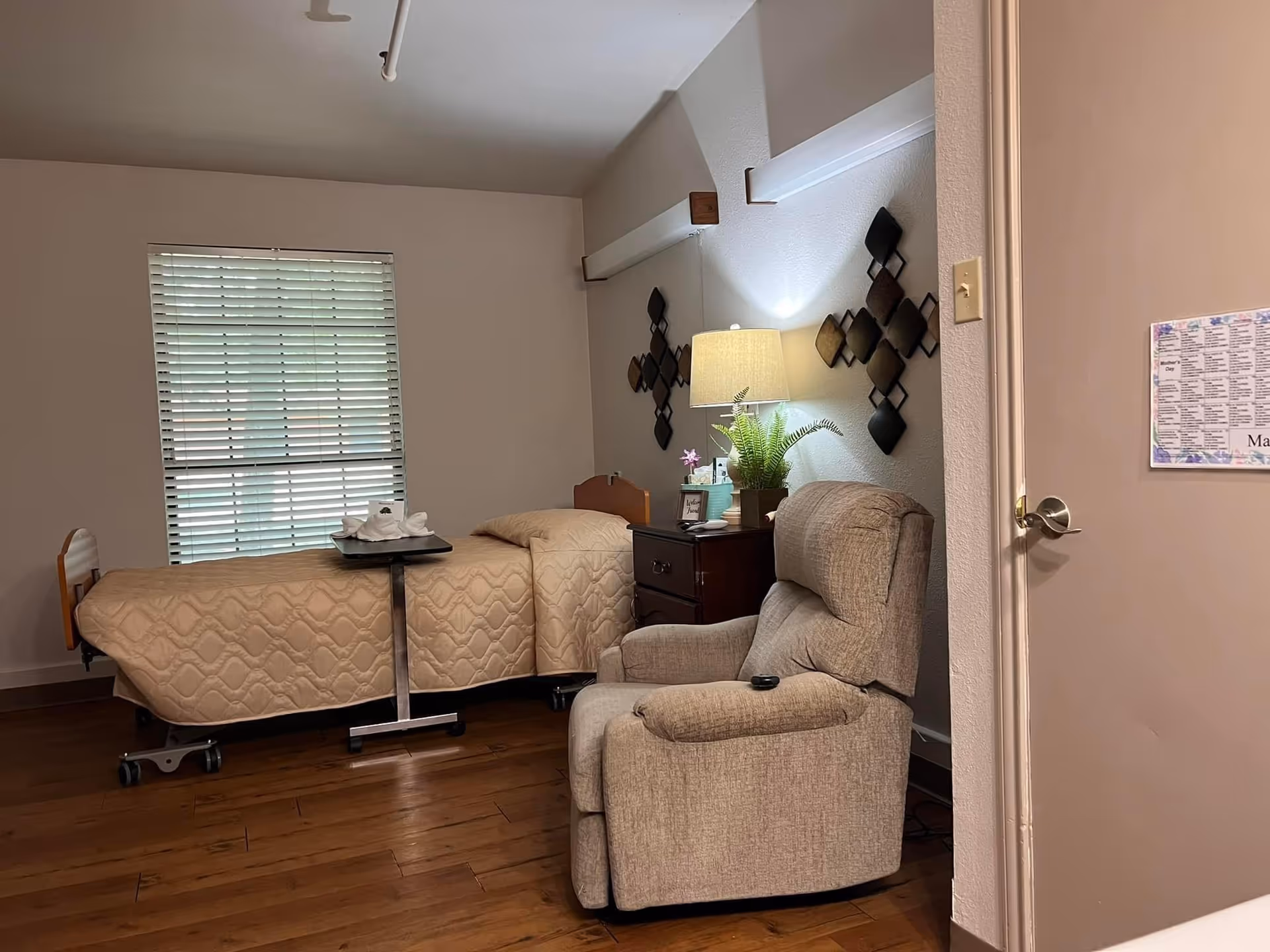 A cozy nursing facility bedroom with a single bed covered in a beige quilt, a small overbed table with folded towels, a wooden nightstand with a lamp, a potted plant, and decorative wall art. There is a comfortable beige recliner chair in the foreground and a window with closed blinds in the background.