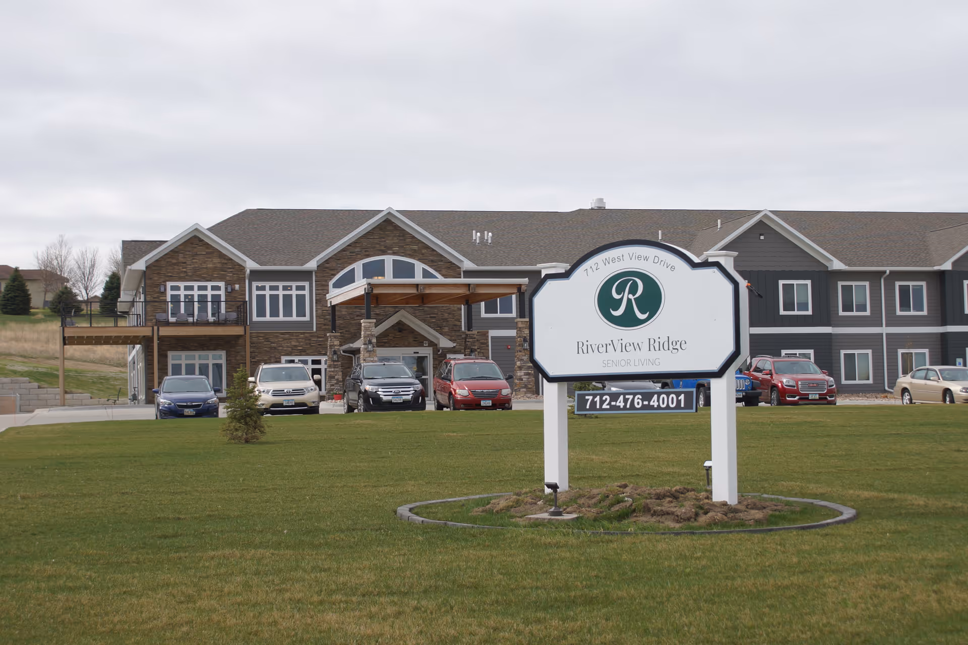 Exterior view of the RiverView Ridge senior living facility with a large sign in the foreground displaying the facility's name, logo, address, and phone number. Several cars are parked in front of the building, which features a combination of stone and siding on its facade, multiple windows, and a covered entrance.