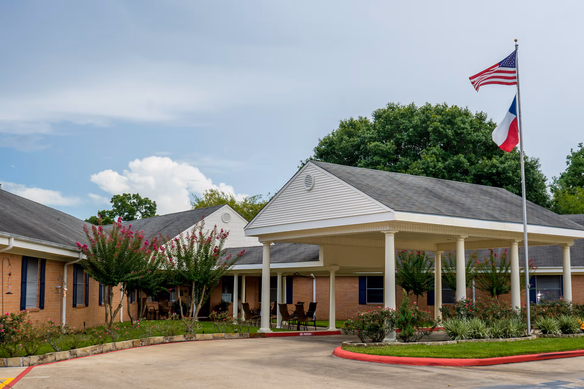 Front entrance of a single-story brick senior care building with a covered portico, U.S. and state flags, and landscaped grounds.