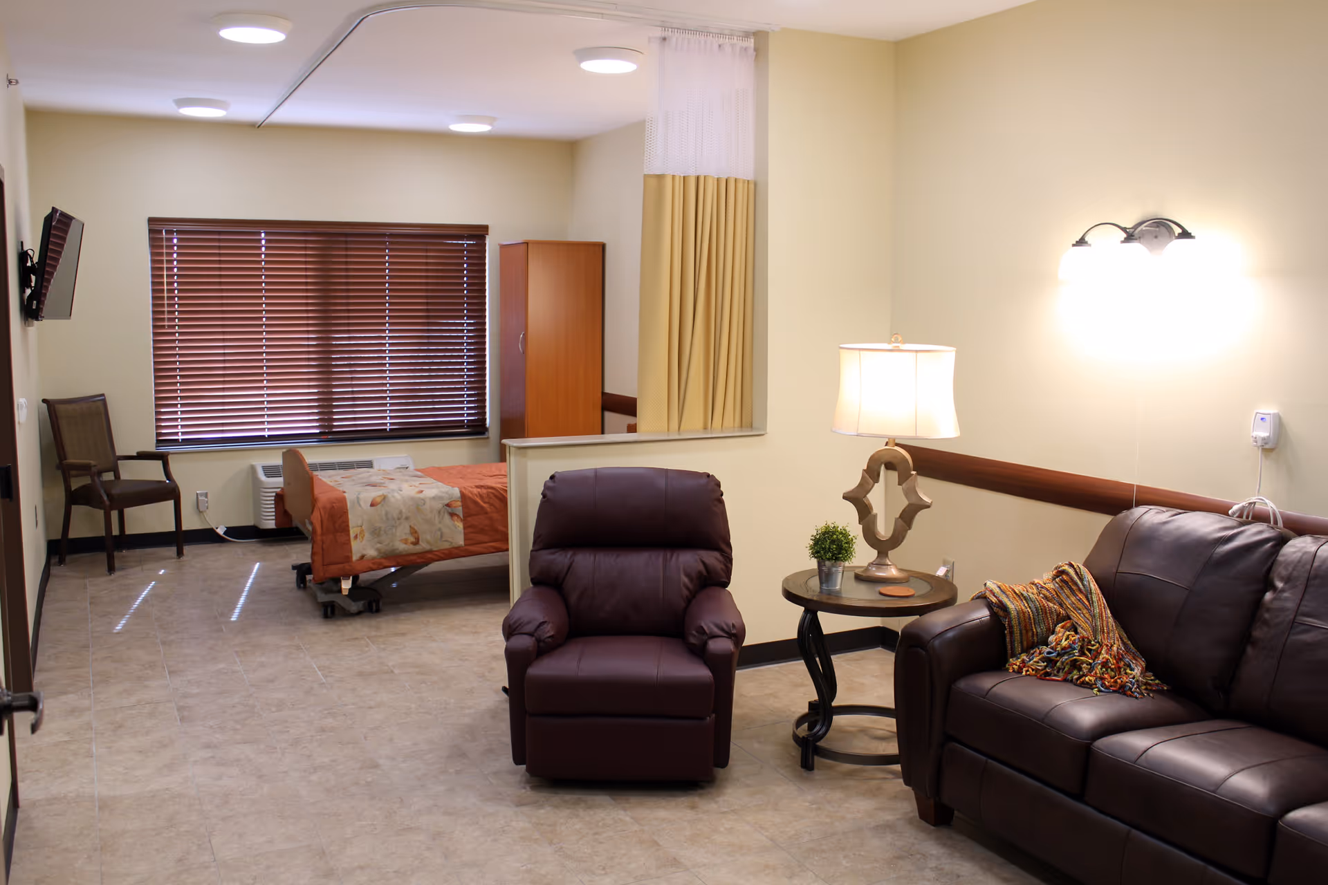 Senior living suite with sofa, recliner, side table, lamp, and a bed behind a divider.