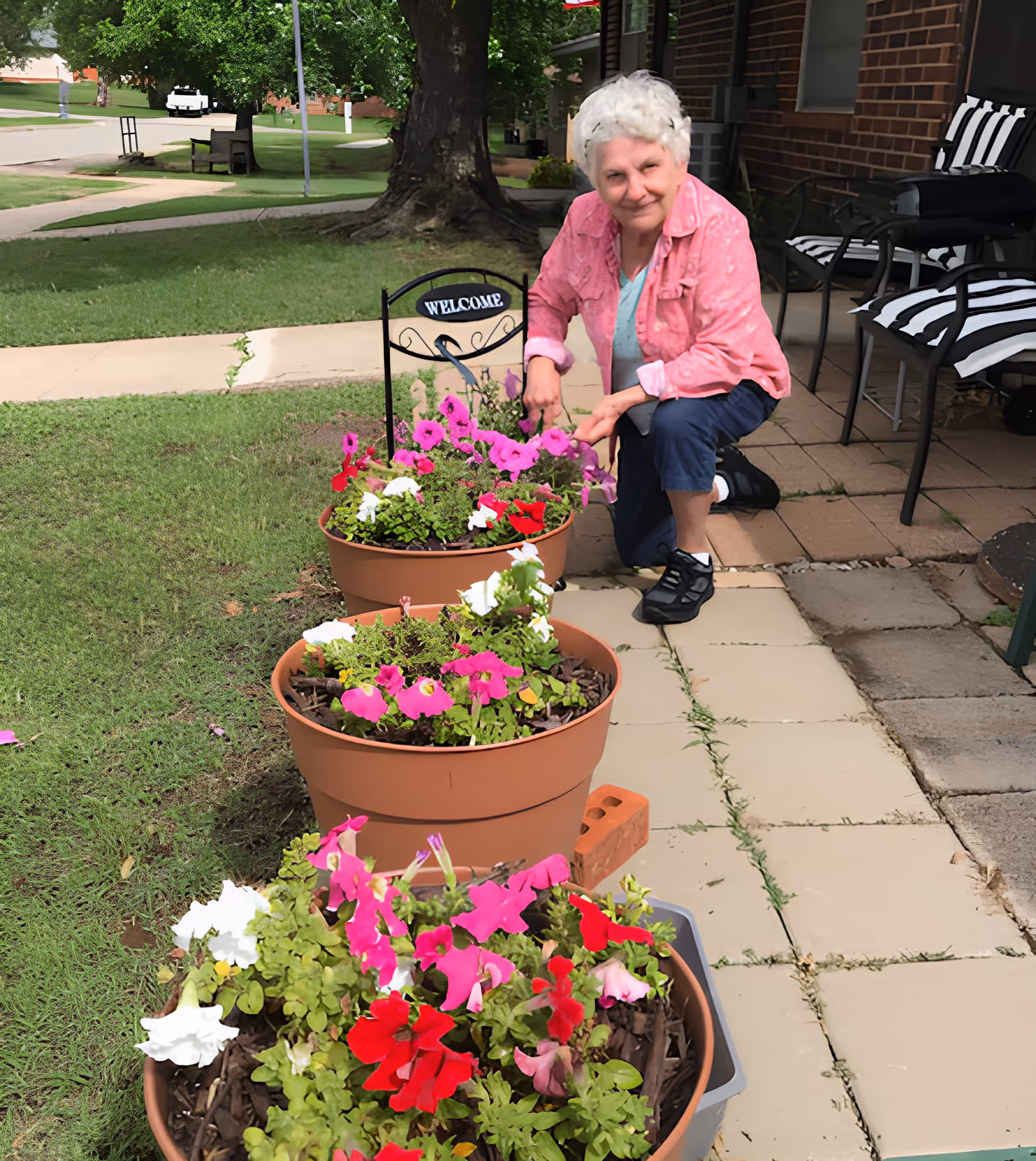An elderly woman kneels on a patio tending several large pots of colorful flowers outside a brick building.