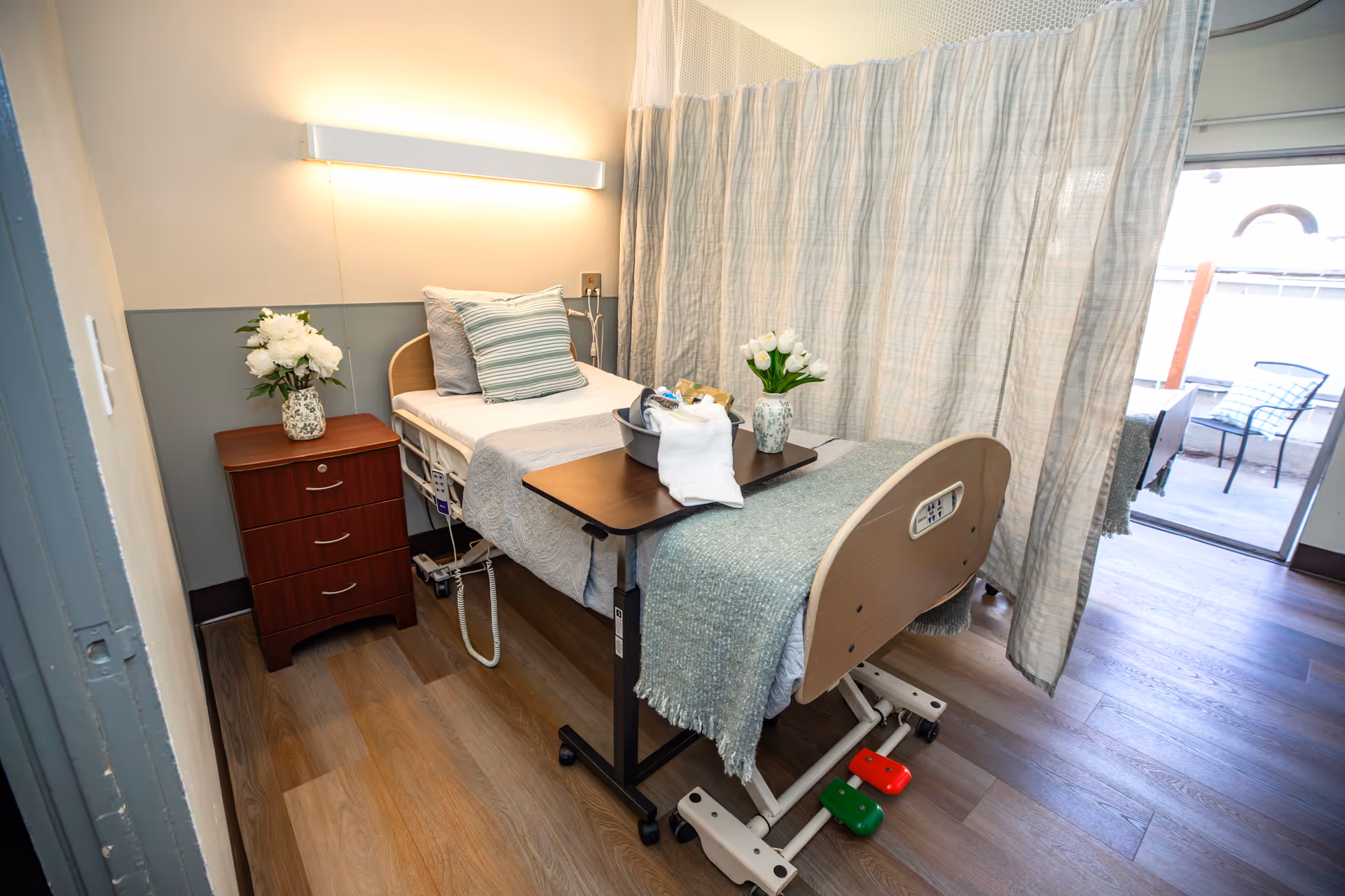 Senior living bedroom with hospital-style bed, privacy curtain, nightstand, and balcony view.