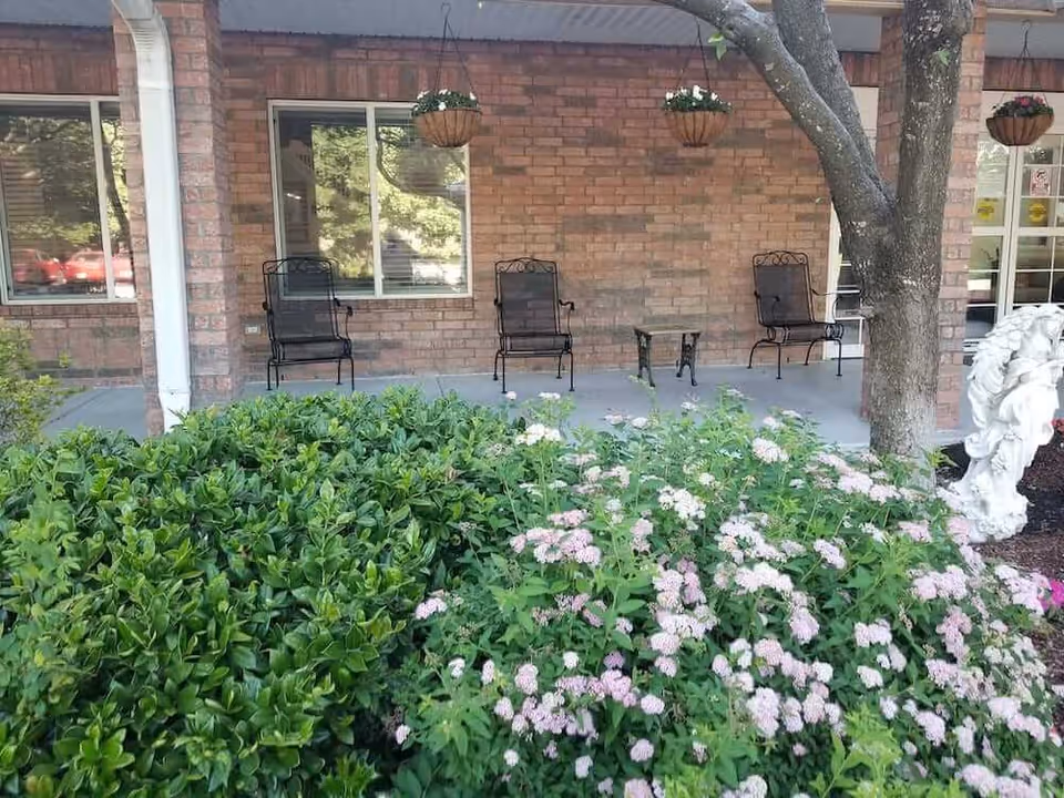 Front porch of a brick building with metal chairs, hanging planters, a tree and flowering shrubs in the foreground.