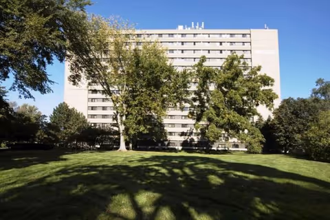 Tall white senior living building behind a grassy lawn and leafy trees.