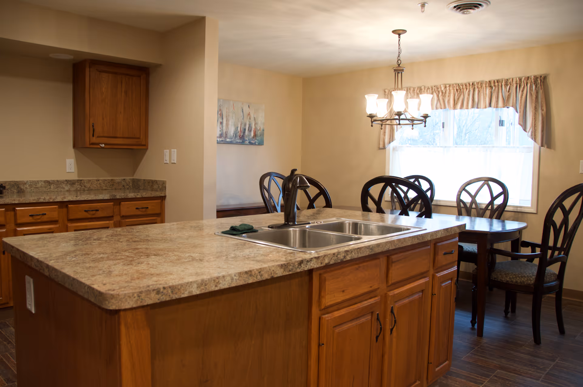 A kitchen and dining area with a large island countertop featuring a double sink and faucet. The kitchen has wooden cabinets and drawers with a marble-like countertop. In the background, there is a dining table with six chairs near a window with curtains and a chandelier overhead. The walls are painted beige and the floor has dark wood-like tiles.