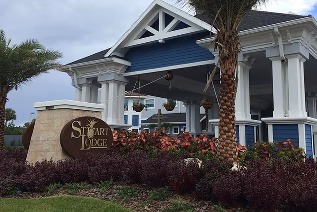 Entrance area of a facility named Stuart Lodge featuring a covered driveway with white pillars and blue siding, surrounded by landscaped plants and palm trees under a cloudy sky.