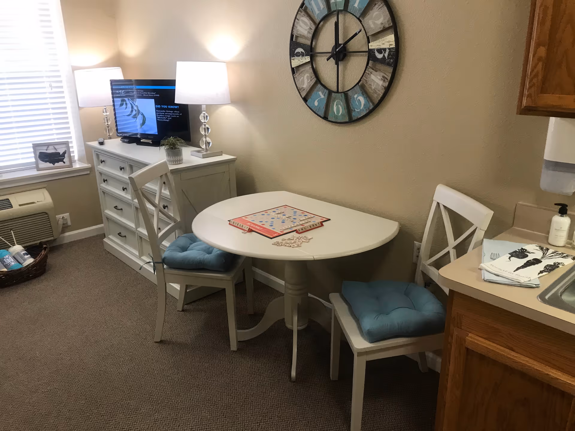 Cozy senior living dining nook with round table, two cushioned chairs, clock, and TV.