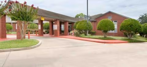 Brick facility entrance with a covered drive-through, manicured shrubs, and a paved parking area.