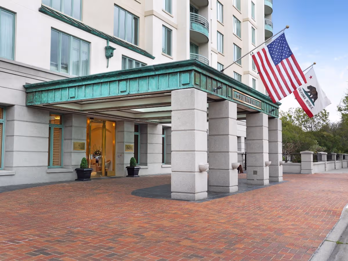 Entrance of The Stratford facility featuring a covered driveway with stone pillars, two flags (American and California Republic) mounted on the right side, and glass doors leading inside. The building exterior is light-colored with multiple windows.