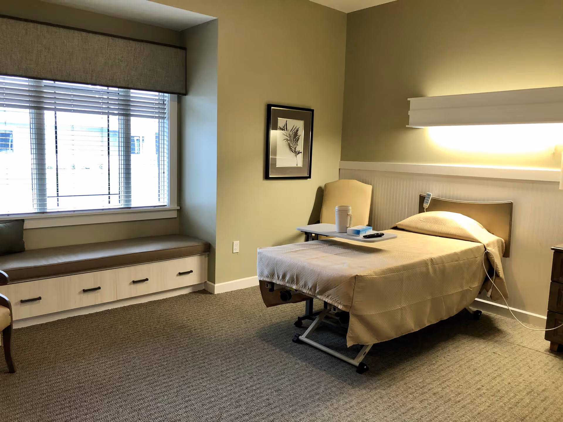 A well-lit bedroom in a senior living facility with a single hospital-style bed covered in beige bedding. There is a small overbed table with a cup and remote control on it. Next to the bed is a beige armchair and a framed botanical picture on the wall. A window seat with cushions is positioned under a large window with blinds. The room has beige walls and carpeted flooring.