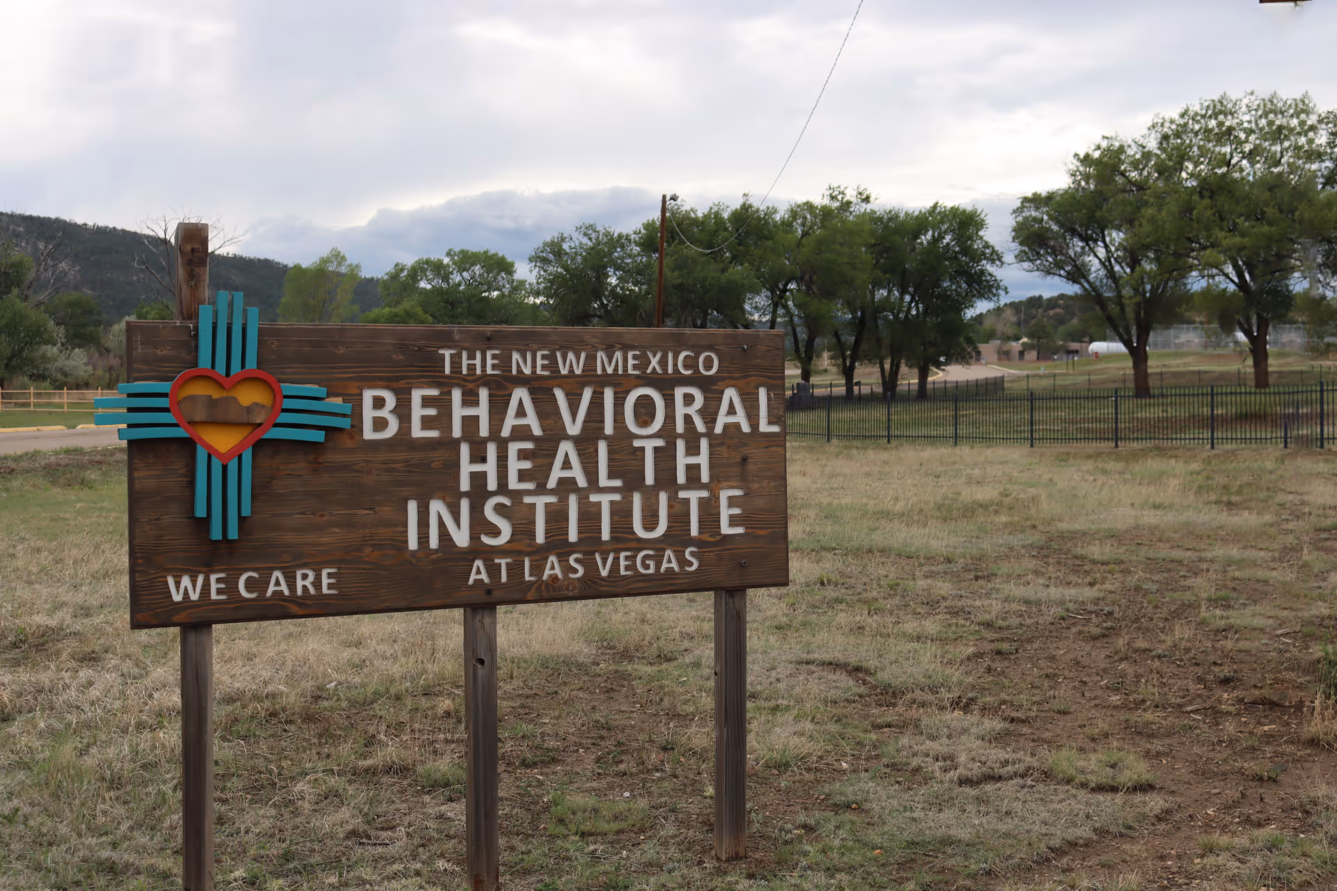 A wooden sign outdoors that reads 'The New Mexico Behavioral Health Institute at Las Vegas' with a colorful heart and cross symbol on the left side, set in a grassy area with trees and a fence in the background under a cloudy sky.