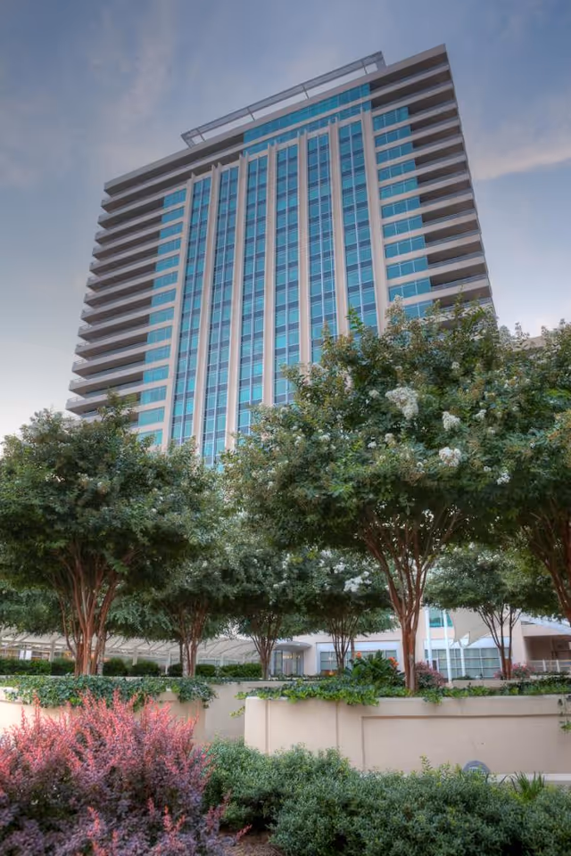 Tall modern high-rise building with blue-tinted glass windows rising above landscaped trees and shrubs in the foreground.