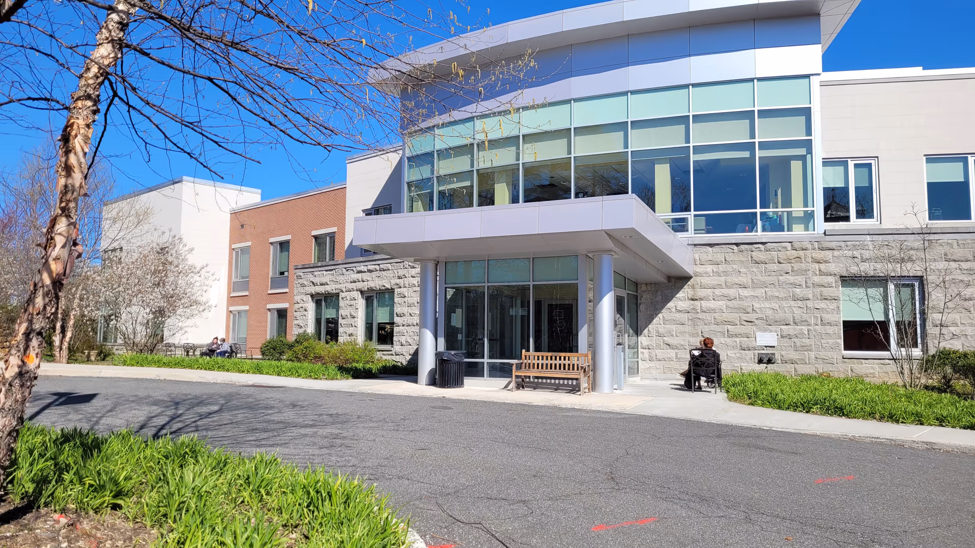 Modern senior living facility exterior with glass entrance, bench, and a person in a wheelchair.