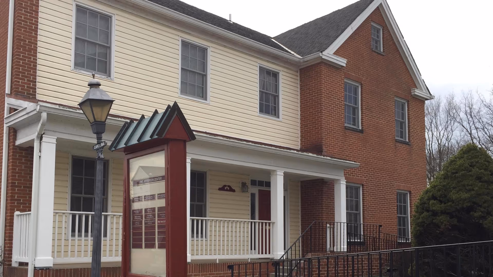 Two-story brick and beige-siding building with a front porch, railing, and a signboard.