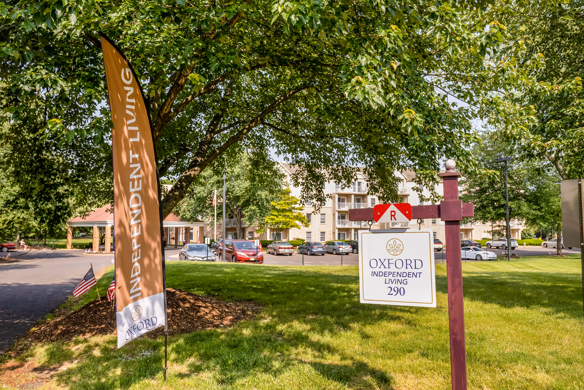 Outdoor view of Oxford Enhanced Senior Living facility showing a grassy area with trees, a signpost with the Oxford Independent Living logo and address number 290, a tall flag with the words Independent Living, and a parking lot with cars and a multi-story building in the background.
