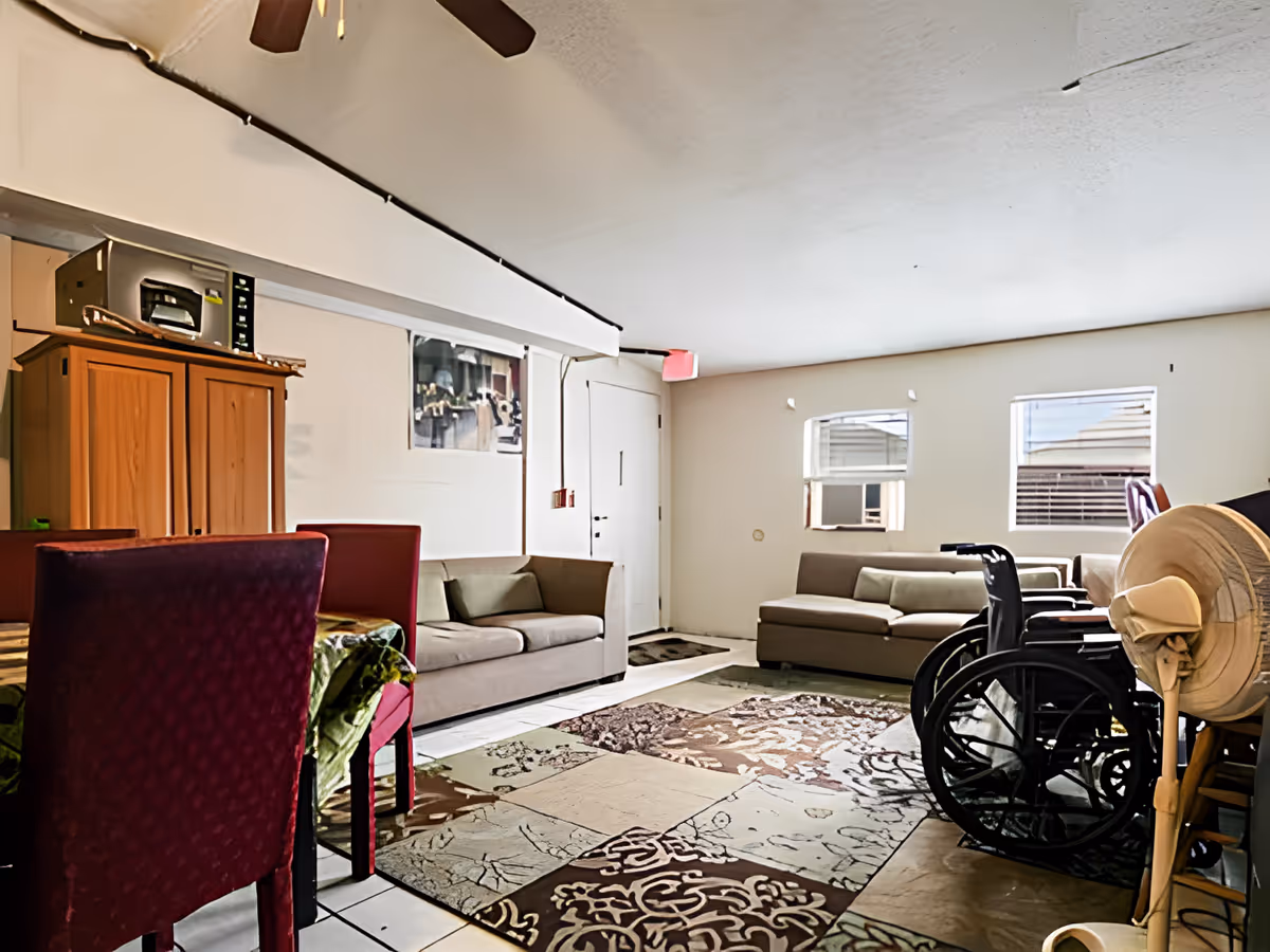 Interior view of a living room area with two beige sofas, a patterned area rug, a wheelchair, a standing fan, and a dining table with red chairs. There is a ceiling fan above and two windows letting in natural light.