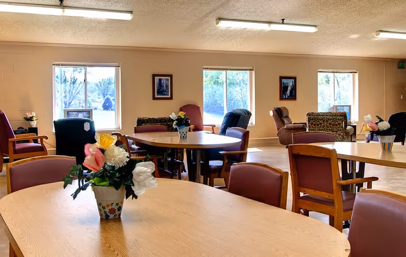 A bright and spacious common area with several round wooden tables and chairs arranged around the room. Each table has a small floral arrangement in a decorative pot. The room has large windows letting in natural light, beige walls, and comfortable armchairs placed near the windows. Framed pictures hang on the walls.