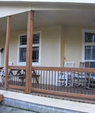 Covered porch with wooden railing and chairs along a beige exterior wall.