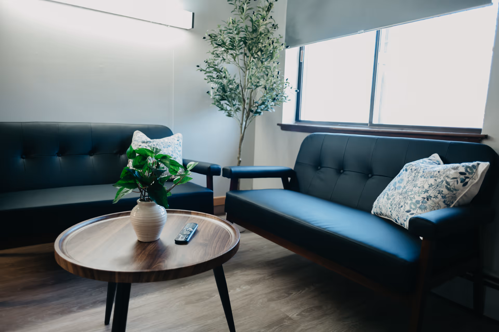 Two black leather sofas, a round wooden coffee table, and a plant in a bright lounge.