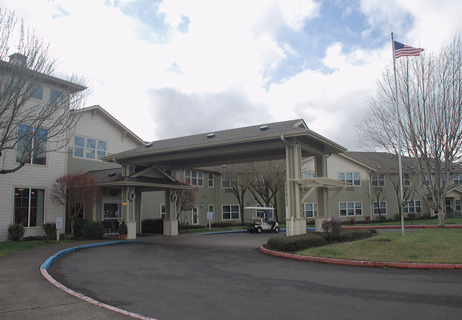 Front entrance of a two-story senior living facility with a covered porte-cochère, circular driveway, flagpole, and a parked golf cart.
