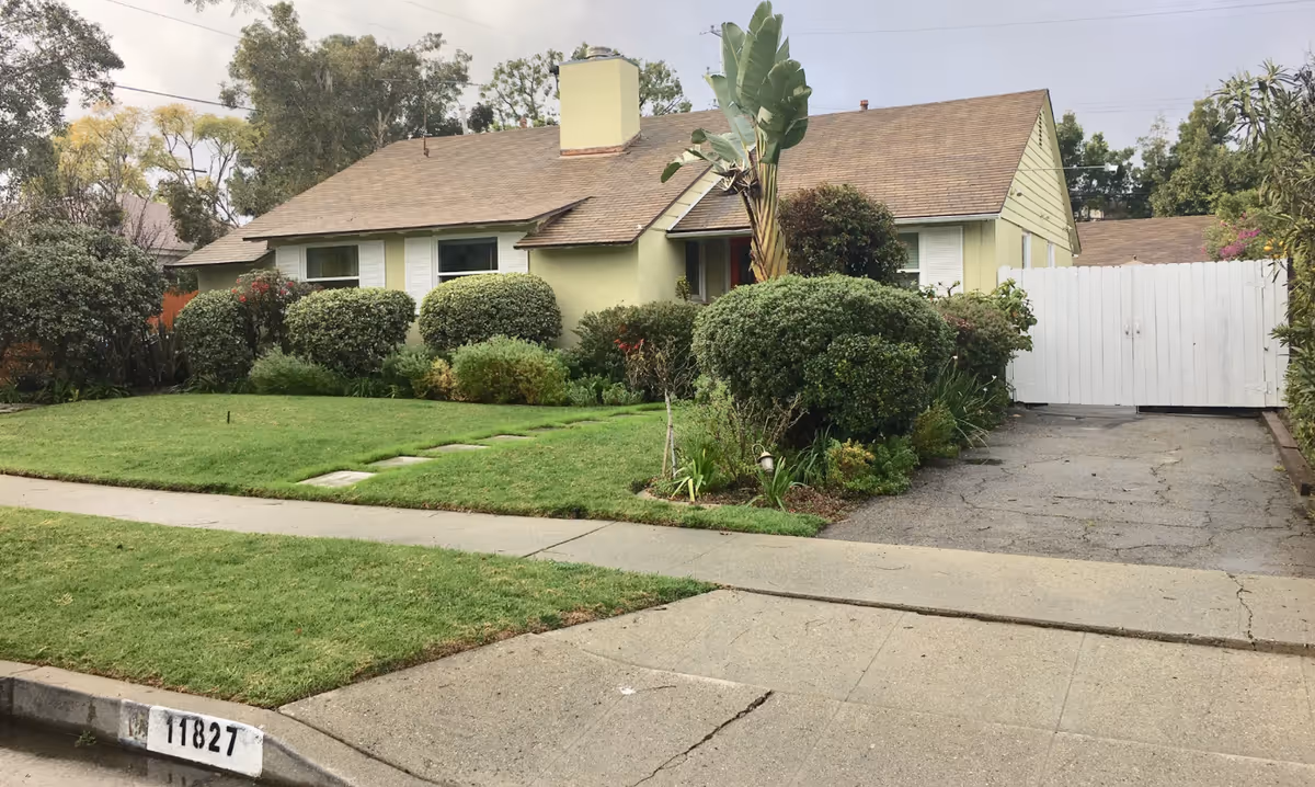 Single-story light-green house with manicured front yard, trimmed shrubs, and a white gate.