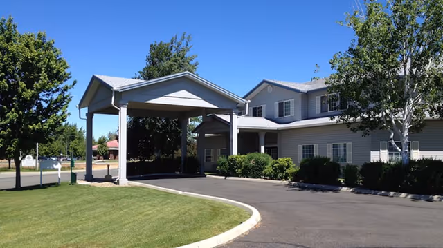 Exterior view of a senior living facility building with a covered entrance driveway, surrounded by green trees and bushes under a clear blue sky.